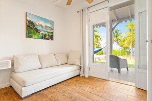 Een kamer met een witte bank, een schilderij van een tropisch landschap en uitzicht op palmbomen door een glazen deur.