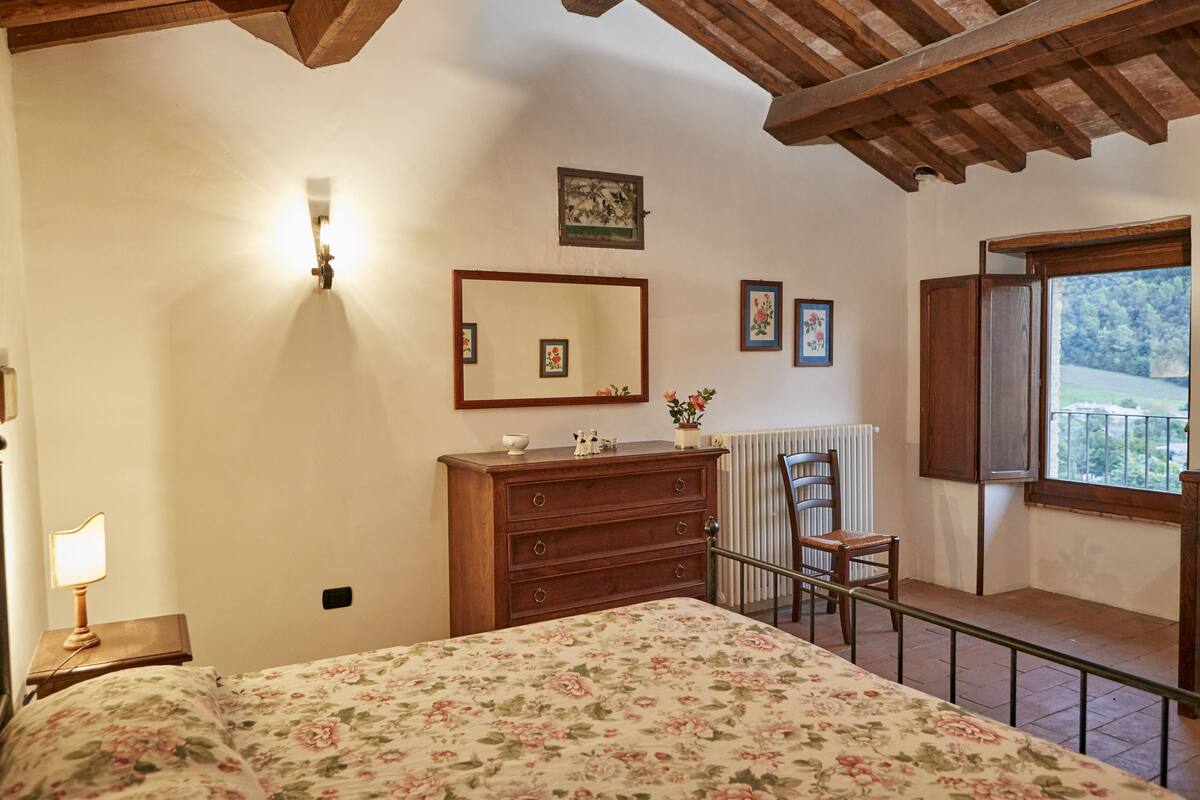 Ein Schlafzimmer mit Bett, Kommode, Spiegel, Heizkörper, Sessel und einem Fenster mit Blick.