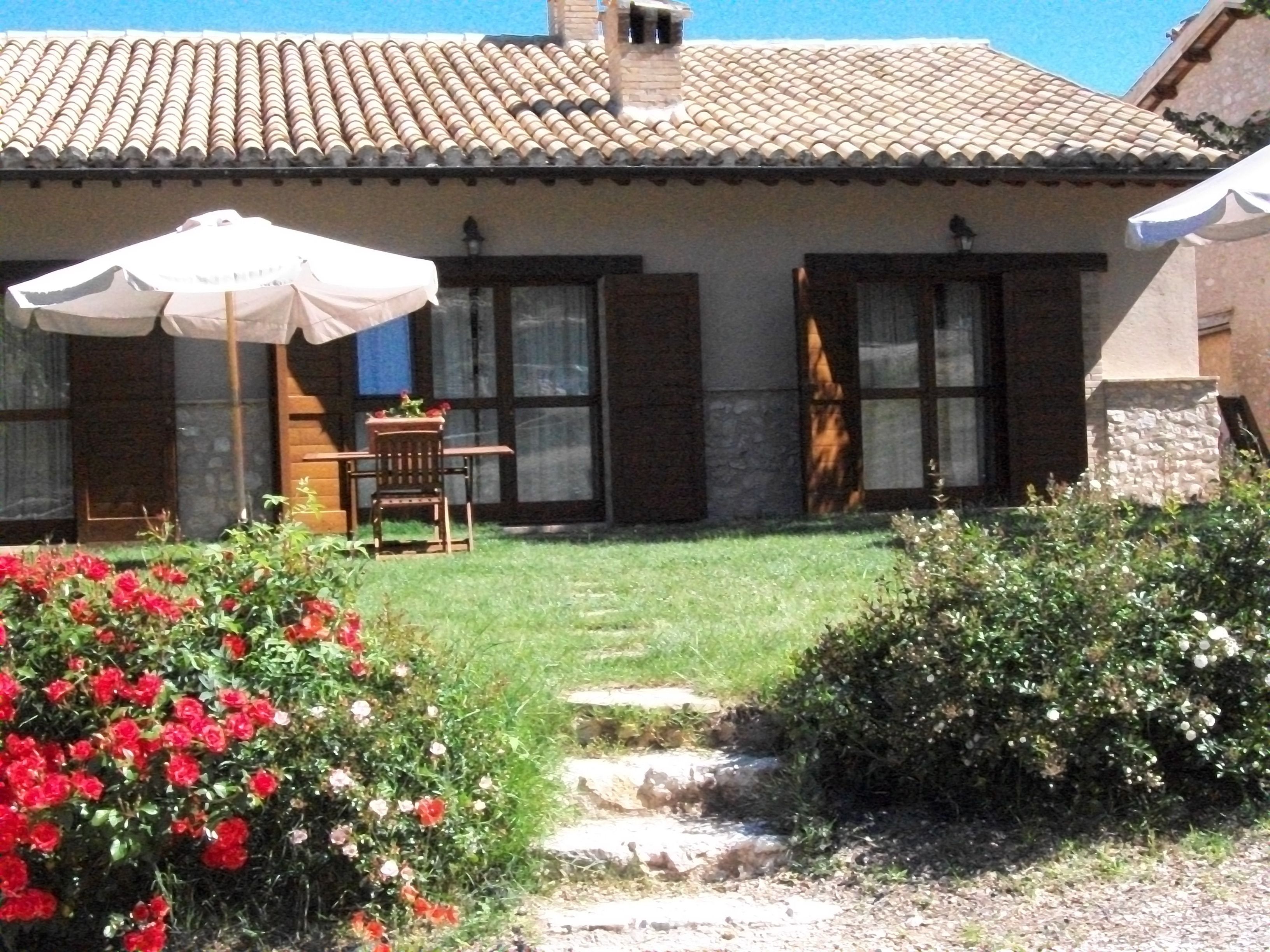 Una casa con tetto in tegole, un patio con tavolo e sedie e un giardino con fiori rossi.