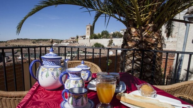 A table set with a pitcher, teapot, cups, and a glass of orange juice, with a view of a historic town and a palm tree. - Hostal Chinchon (Chinchon)