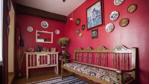 A room with a red wall, a wooden bench with a floral cushion, a mirror, and various decorative plates and coins on the wall. - Hostal Chinchon (Chinchon)