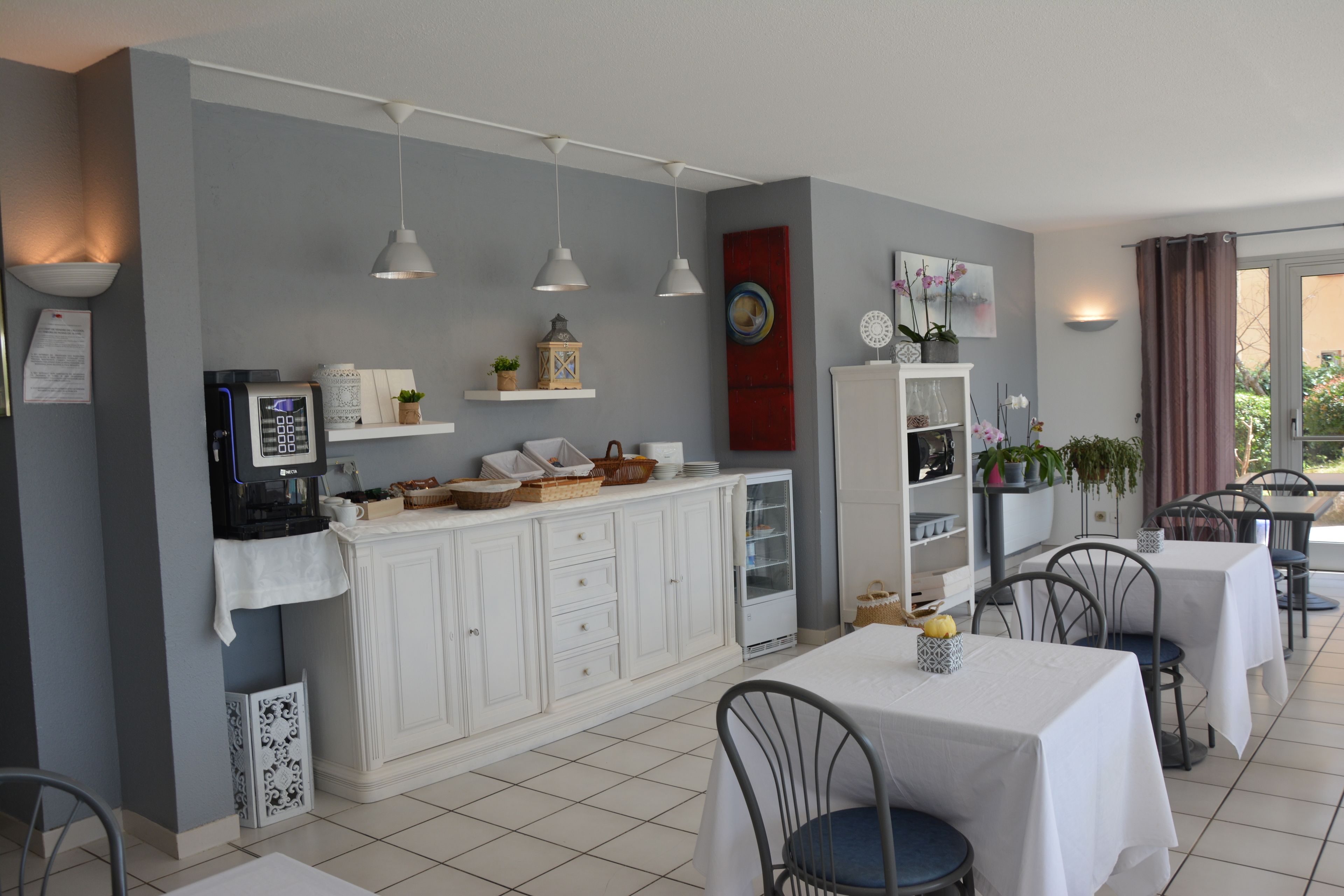 Un espace repas moderne avec des tables et des chaises blanches, une machine à café et des objets décoratifs disposés sur des étagères.