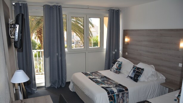 Habitación de hotel con dos camas, un televisor, una lámpara y vista a palmeras a través de la ventana.