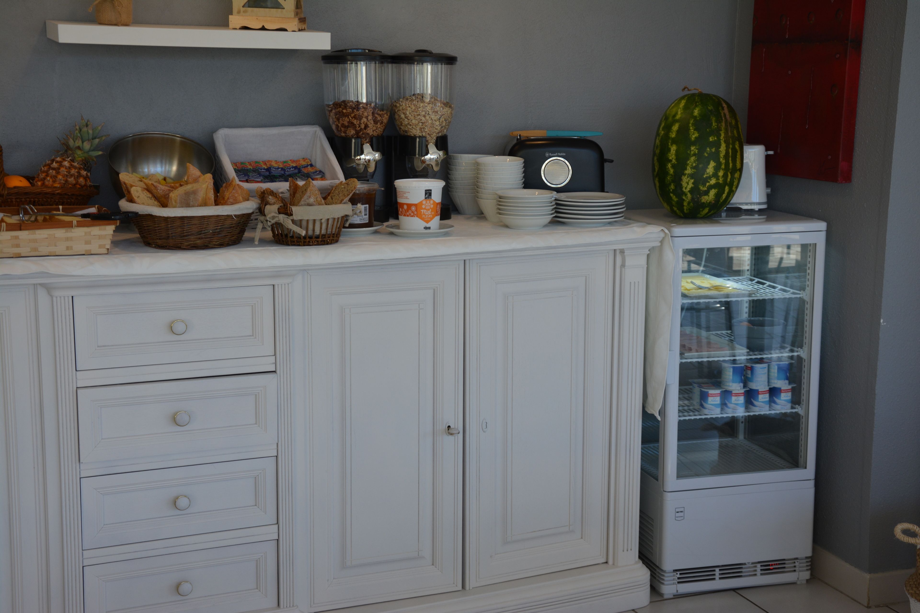 A kitchenette with a white cabinet, a small refrigerator, and various food items like bread and a watermelon.