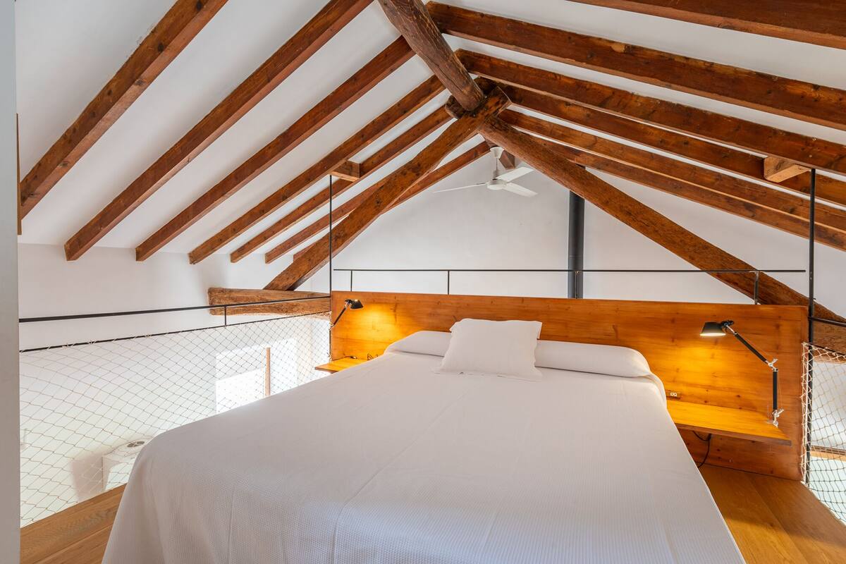A bedroom with a sloped ceiling, wooden beams, a bed with white bedding, a wooden headboard, and a ceiling fan.