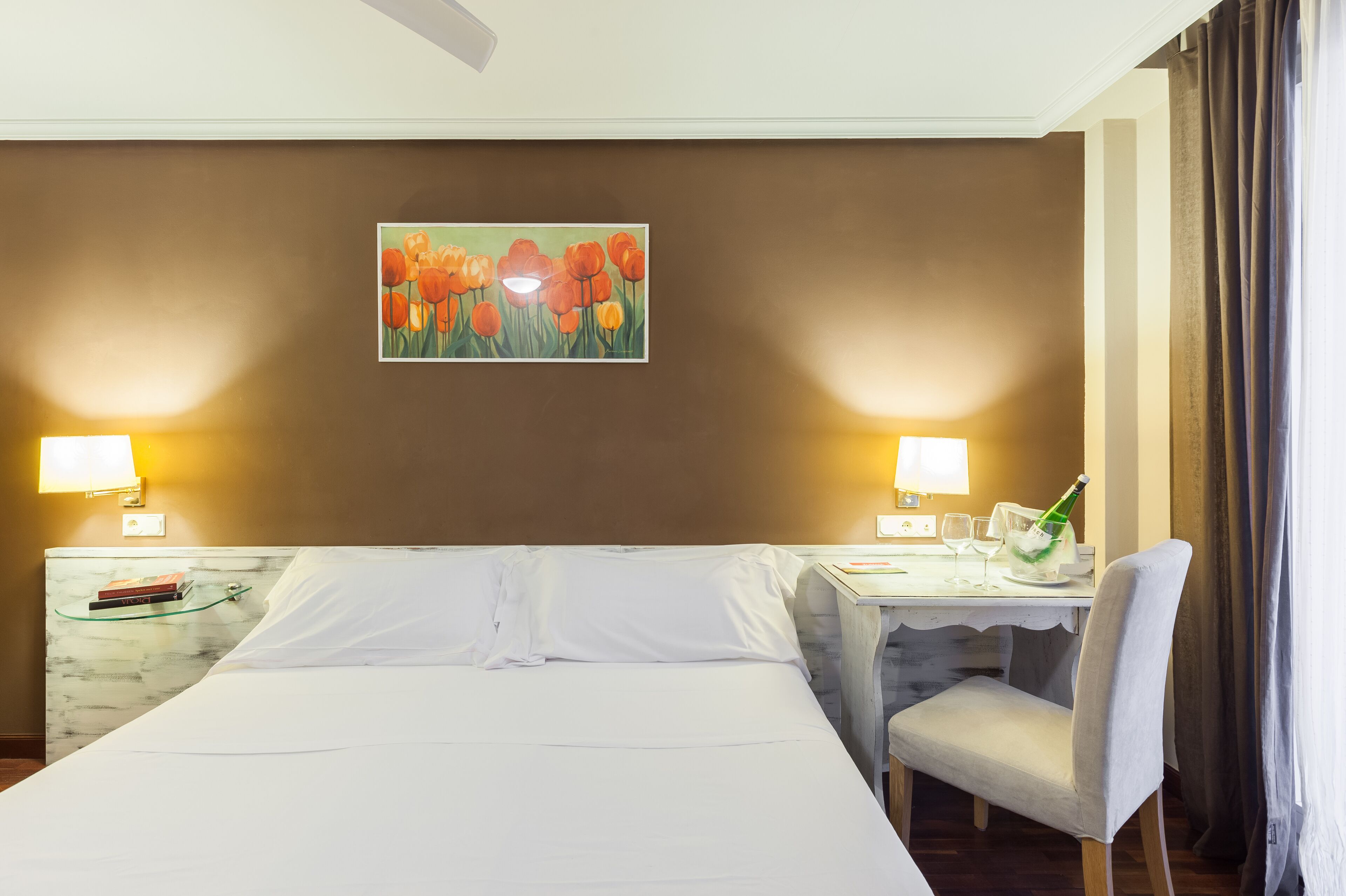 A hotel room with a large bed, two bedside tables with lamps, a small desk with a chair, and a painting of tulips on the wall.
