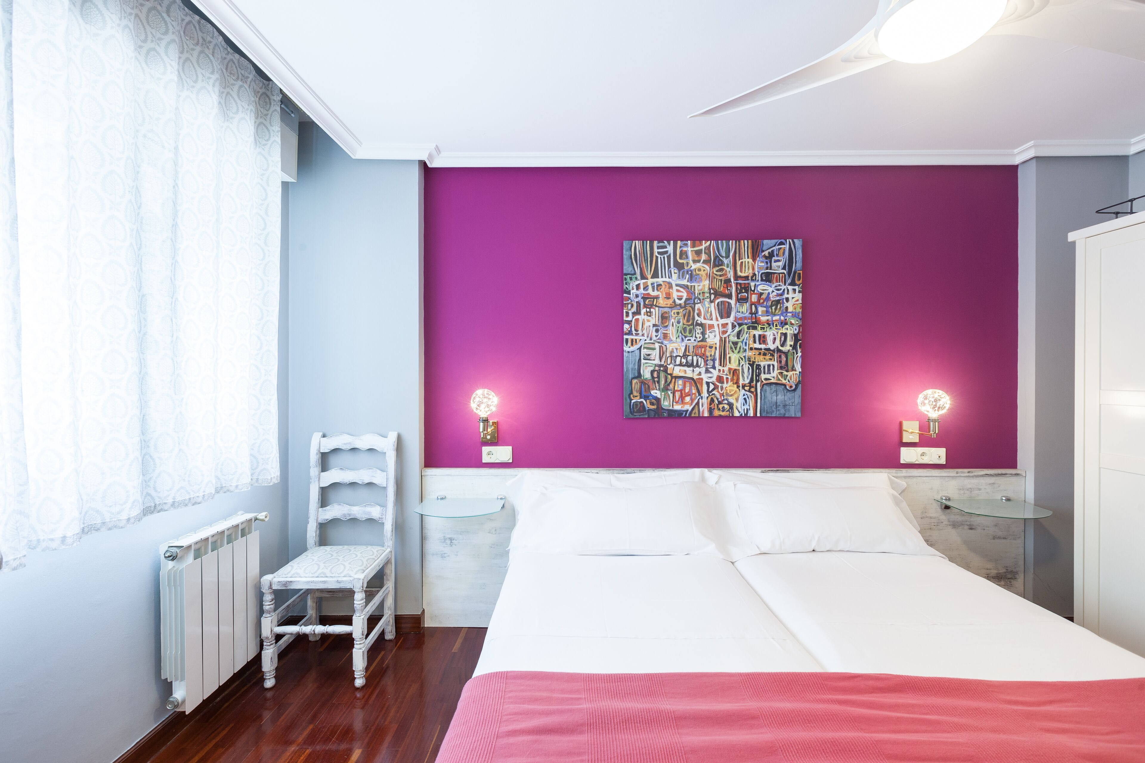 A bedroom with a bed, a chair, a radiator, a wall with a colorful artwork, and two bedside lamps.