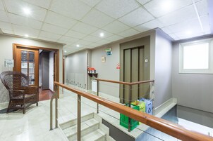Un espacio interior limpio y bien iluminado con una silla de madera, una escalera con barandilla metálica y un contenedor verde para reciclaje.