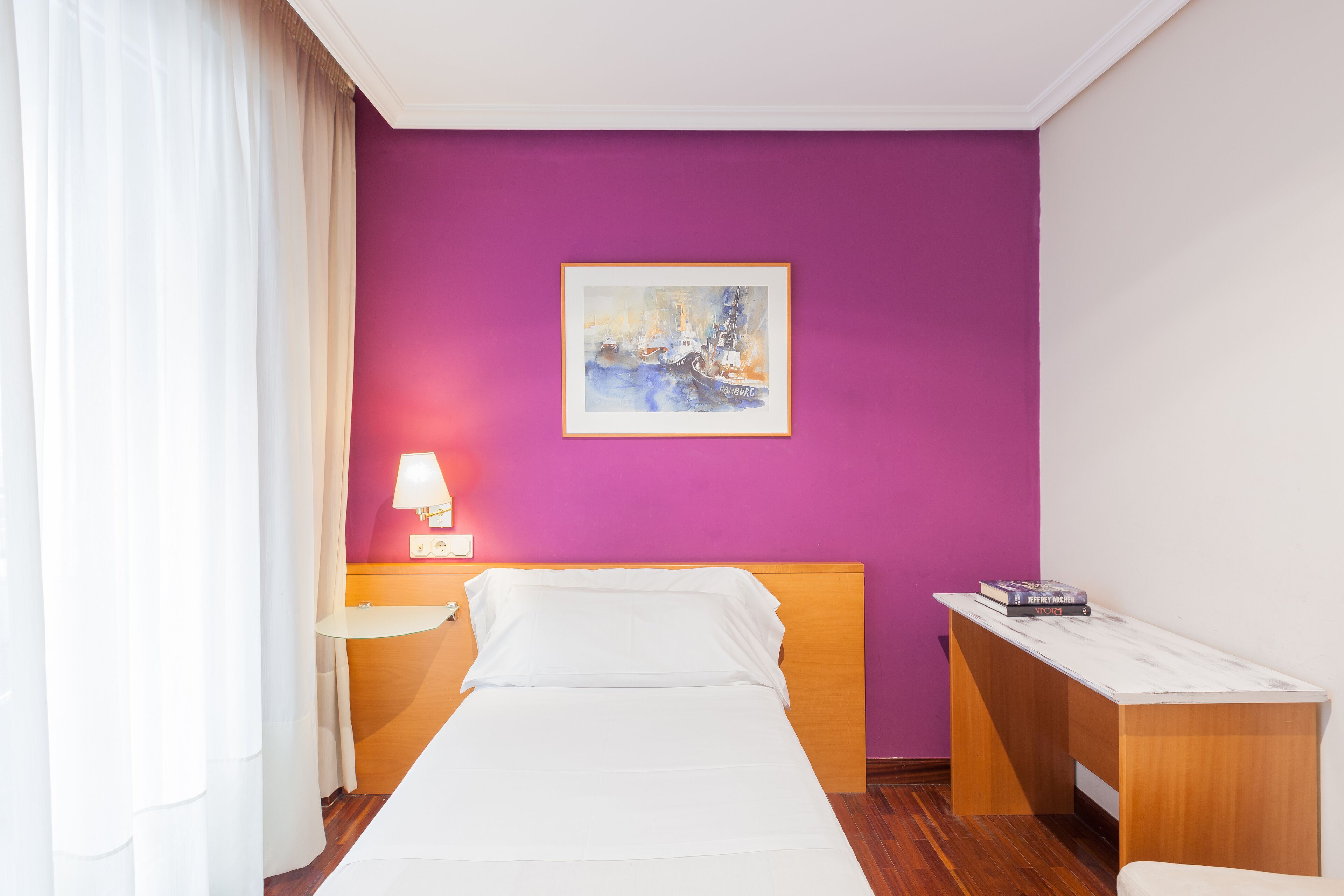 A hotel room with a bed, a nightstand, a desk, a lamp, and a painting on the wall.