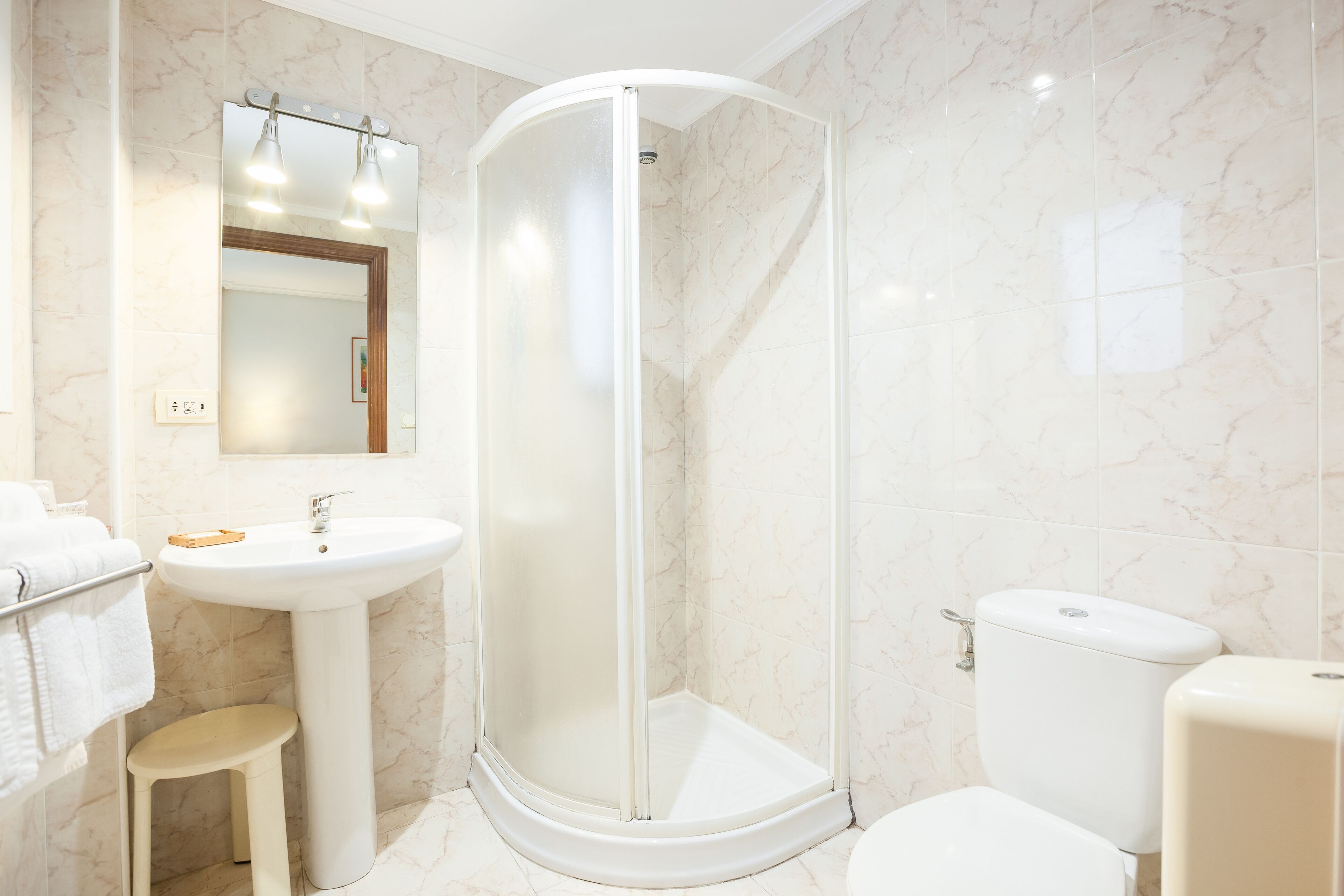 A bathroom with a shower enclosure, sink, toilet, and a small stool.
