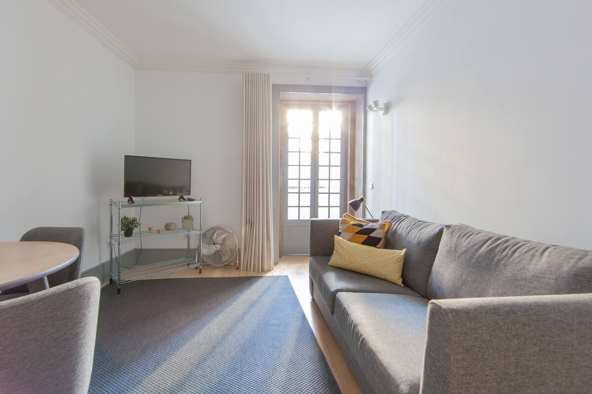 A living room with a grey sofa, a TV on a metal stand, a small table, and a window with curtains.