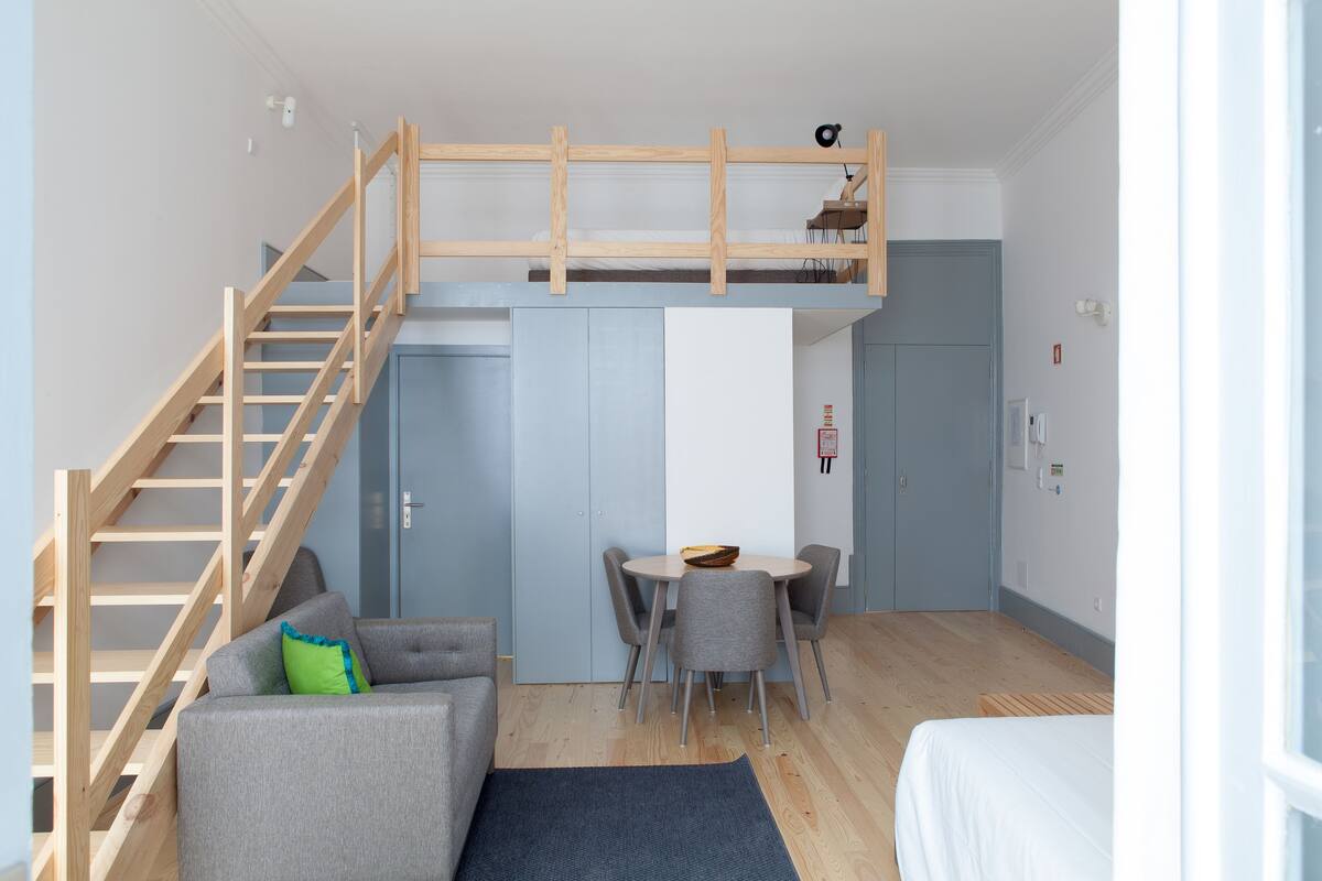 A compact room with a loft bed, a built-in desk, a dining table, and a staircase.