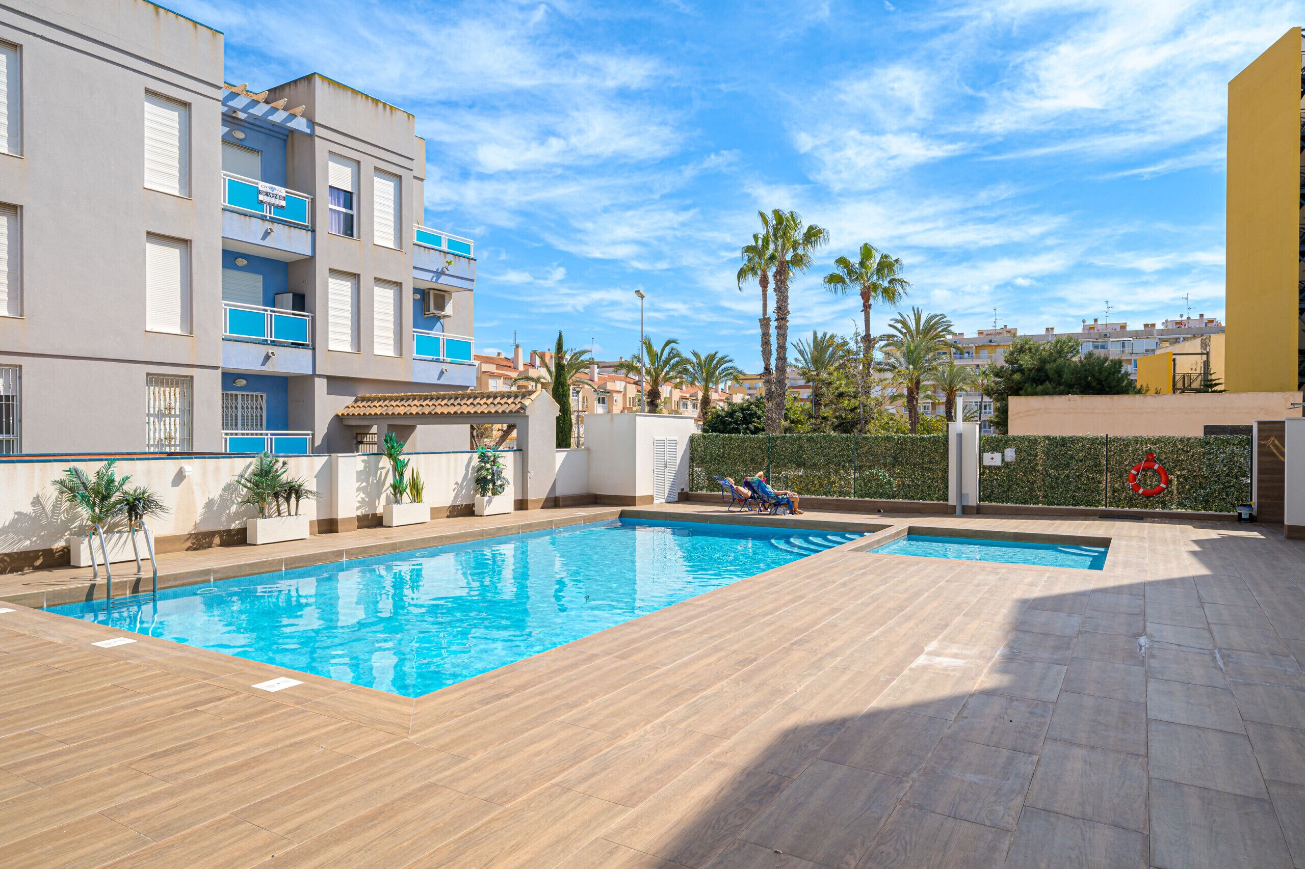 Une piscine avec deux chaises longues, une bouée de sauvetage et une vue sur un quartier résidentiel bordé de palmiers.