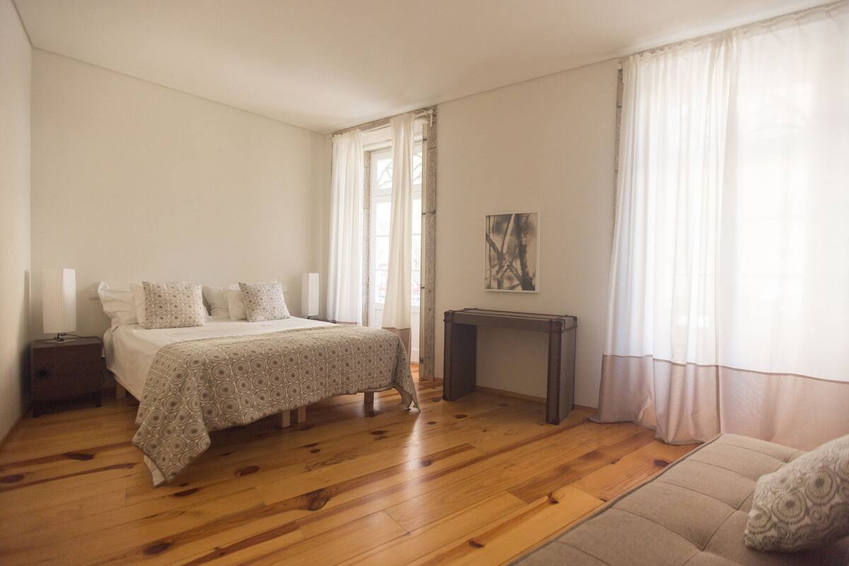A bedroom with a wooden floor, a bed with a patterned bedspread, a bedside table, and a window with curtains.