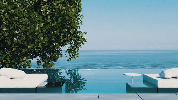 Una piscina infinita con sillones blancos y una mesita, rodeada de exuberante vegetación y vistas despejadas al mar.