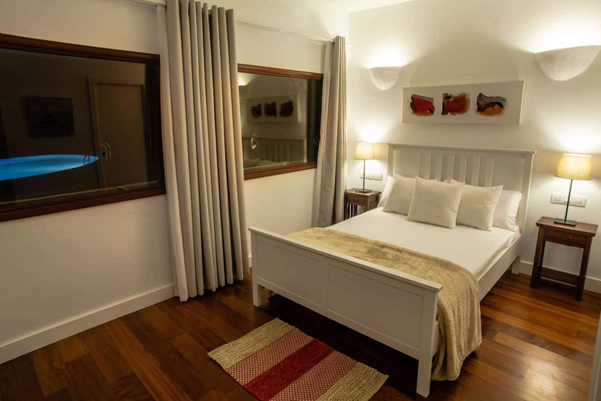 A bedroom with a bed, two bedside tables, a window with curtains, and a wall-mounted artwork.