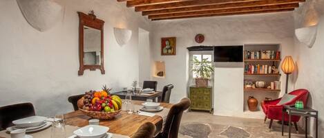 Un comedor con una mesa de madera dispuesta para cuatro personas, rodeada de sillas. Hay un frutero sobre la mesa, una estantería con libros y una silla roja.