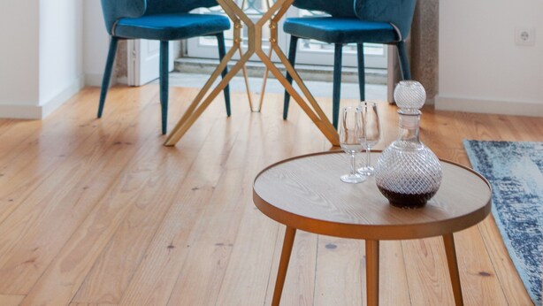 Une pièce dotée d’un plancher en bois, d’une table ronde avec un plateau en verre, de deux chaises bleues, d’une carafe en verre et d’une vue dégagée sur l’extérieur grâce à une grande fenêtre.