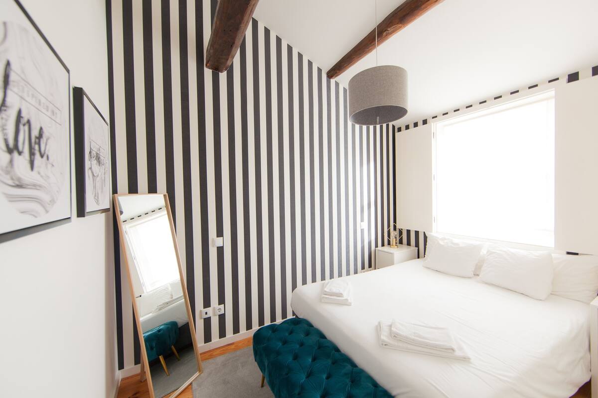 A hotel room with a striped wall, a bed with white linens, a bedside table, a mirror, and a window.