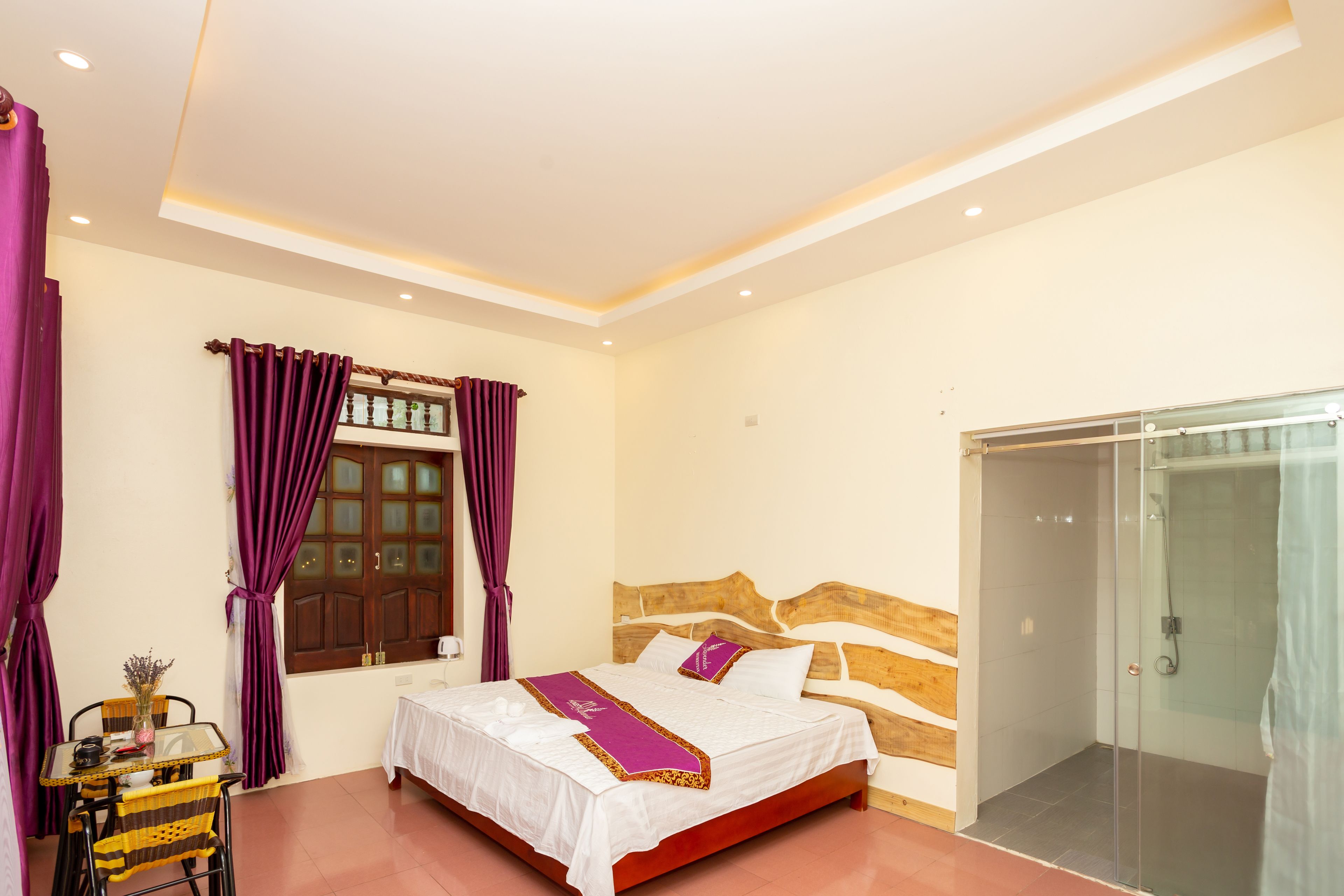 A bedroom with a bed, a glass shower enclosure, a table with chairs, and purple curtains.