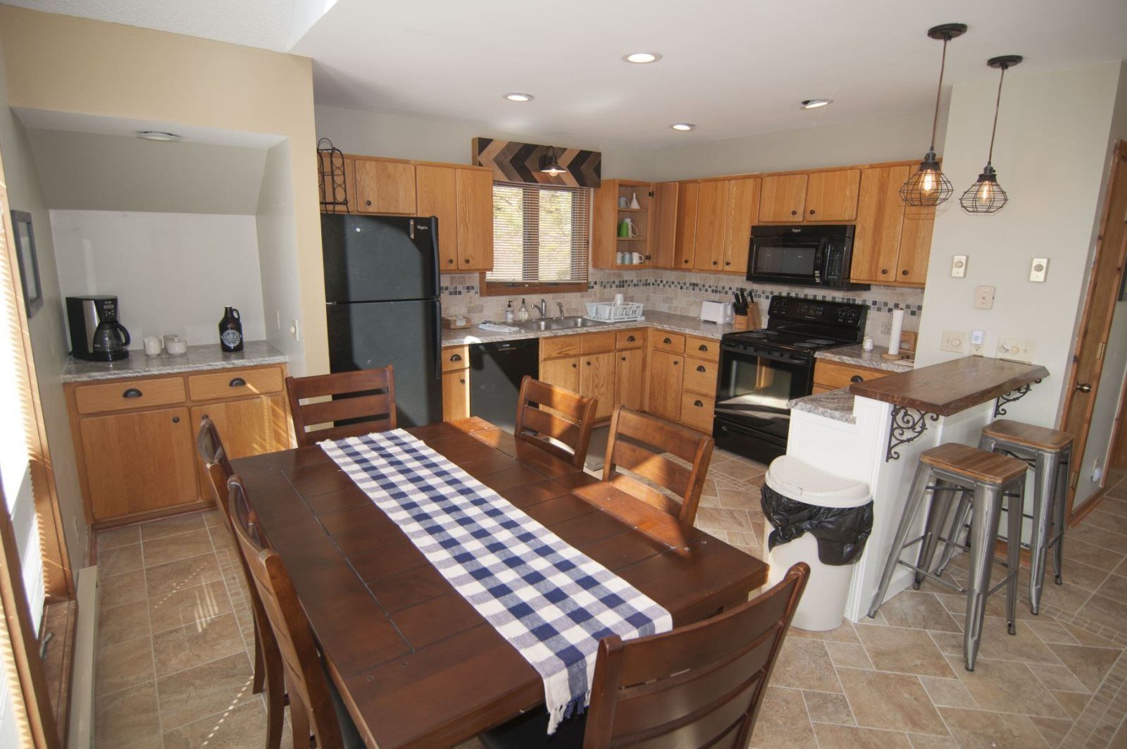 A kitchen with wooden cabinets, a black refrigerator, a microwave, and a stove. There is a dining table with chairs and a checkered tablecloth.