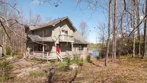 A two-story wooden house with a porch and a red door, surrounded by trees and a body of water. - Woodland Pond (Davis)