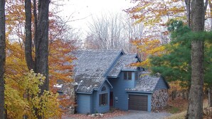 A two-story blue house with a stone foundation, a garage, and a driveway surrounded by trees with autumn leaves.