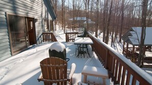 A wooden deck with snow-covered chairs and a table, surrounded by trees and a house.