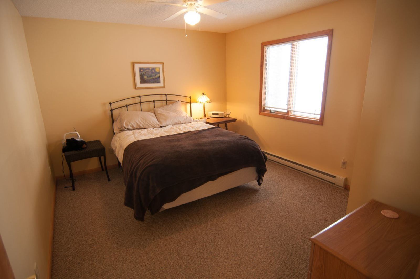 Une chambre avec un lit simple, des tables de chevet, une commode, une lampe, une fenêtre avec des stores et un ventilateur de plafond.