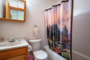A bathroom with a wooden vanity, a toilet, a bathtub, and a shower curtain with a forest scene.