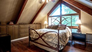 A bedroom with a sloped ceiling, a bed with a patterned bedspread, a nightstand with a lamp, and a window with a view of greenery. - Valley View Lodge (Davis)