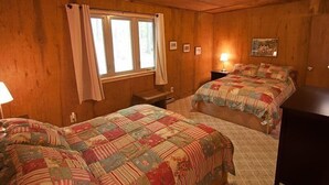 Ett sovrum inrett i stugstil med två sängar, träväggar, ett fönster med gardiner, en lampa och en inramad tavla på väggen.