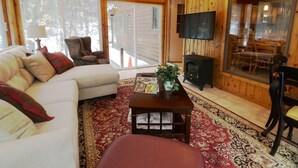 Interior acogedor de una cabaña con sofá, sillón y una mesita con una planta. Un televisor está montado en la pared, y se observa una chimenea al fondo.