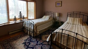 A bedroom with two metal beds, a ceiling fan, a window with blinds, a small Christmas tree, and a patterned rug.