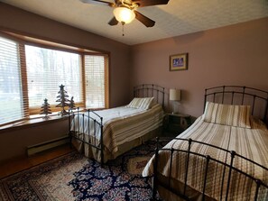 Una camera da letto con due letti in metallo, una ventola a soffitto, una finestra con persiane, un piccolo albero di Natale e un tappeto decorato.