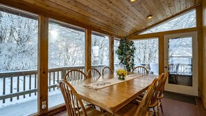 A wooden dining room with a large table and chairs, large windows, and a view of snow-covered trees.
