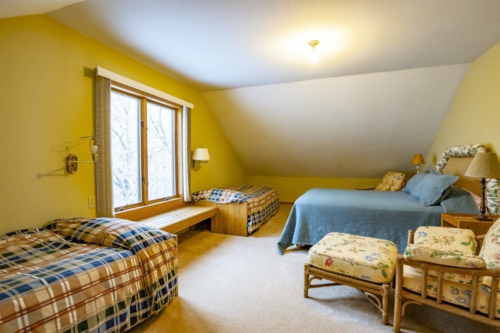 Ein gemütliches Schlafzimmer im Dachgeschoss mit zwei Betten, einer Bank und einem Sessel, mit großem Fenster und Deckenleuchte.