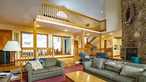 A spacious living room with two sofas, a wooden coffee table, and a stone fireplace. A wooden staircase leads to an upper level with a balcony.
