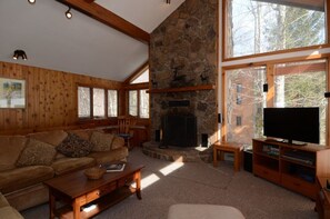 Ein gemütliches Wohnzimmer mit Kamin aus Stein, Holzmöbeln und großen Fenstern mit Blick ins Freie.