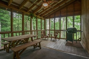 Una terraza de madera con mesas y sillas de picnic, una parrilla y ventanas grandes con vista a un bosque.