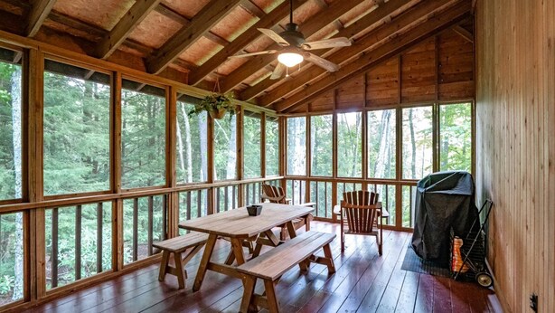 Eine Holzhütte mit einem großen Esstisch und Bänken, einem Deckenventilator und einem Grill vor der Tür.