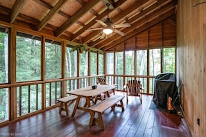 Een houten chalet met een grote eettafel en banken, een plafondventilator en een barbecue buiten.