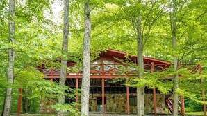 A rustic cabin with a stone foundation, wooden beams, and a red roof, surrounded by lush greenery and trees. - Our Greyt Escape (Davis)