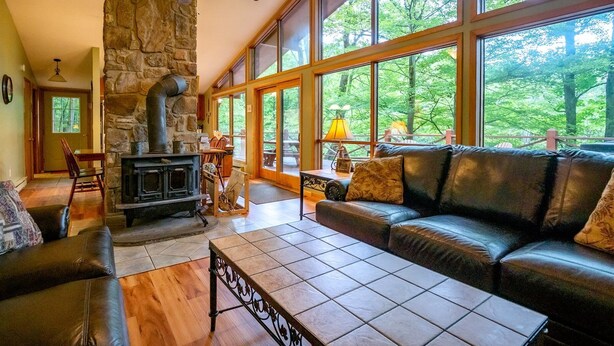 A cozy living room with a stone fireplace, leather sofa, and large windows offering a view of the forest. - Our Greyt Escape (Davis)