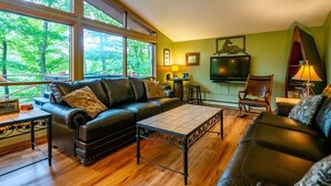 A cozy living room with a leather sofa, a coffee table, and a TV. Large windows offer a view of trees. - Our Greyt Escape (Davis)