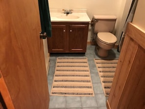 A bathroom with a wooden vanity, a toilet, and two woven mats on the floor. - Northwoods H5 (Davis)
