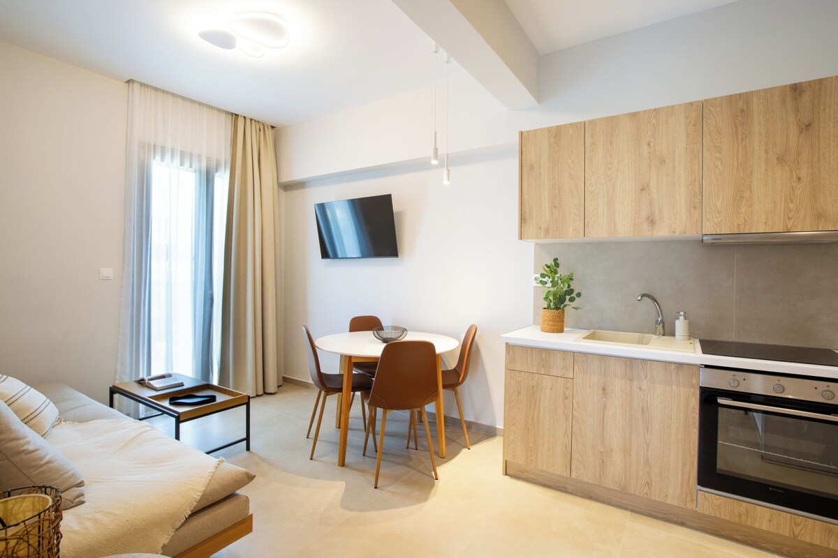 A modern kitchen with wooden cabinets, a dining area with a table and chairs, a sofa, and a flat-screen TV mounted on the wall.