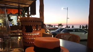 A table with a hat, a bottle, and a glass. There is a view of the beach and cars in the background. - Calmare (Rethymno)