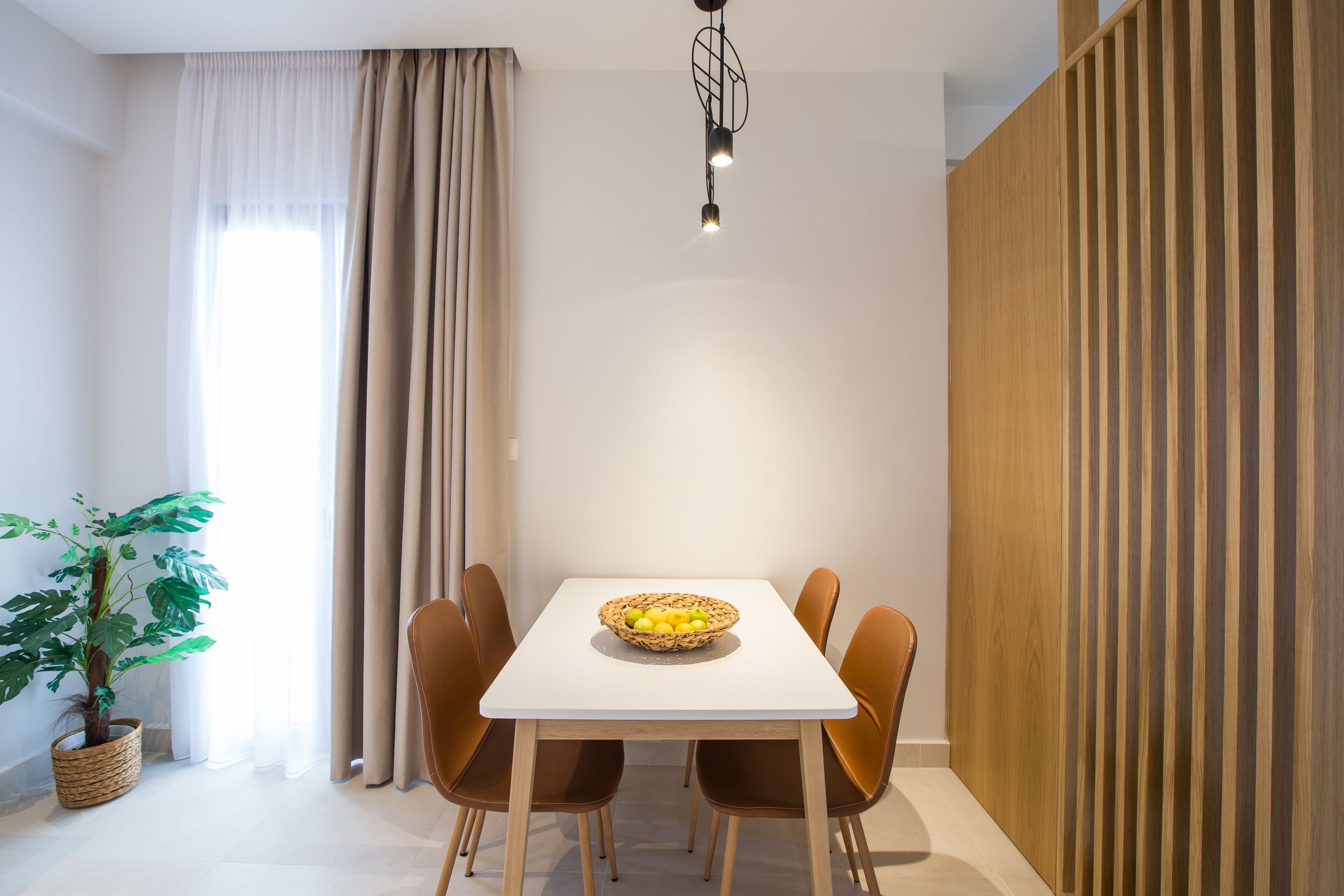 Een eethoek met een witte tafel, zes bruine stoelen, een gevlochten mand met fruit, een potplant en een moderne hanglamp.