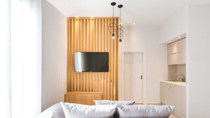 A modern hotel room with a large bed, a flat-screen TV mounted on a wooden panel wall, a kitchen area with white cabinets, and a pendant light fixture. - Calmare (Rethymno)